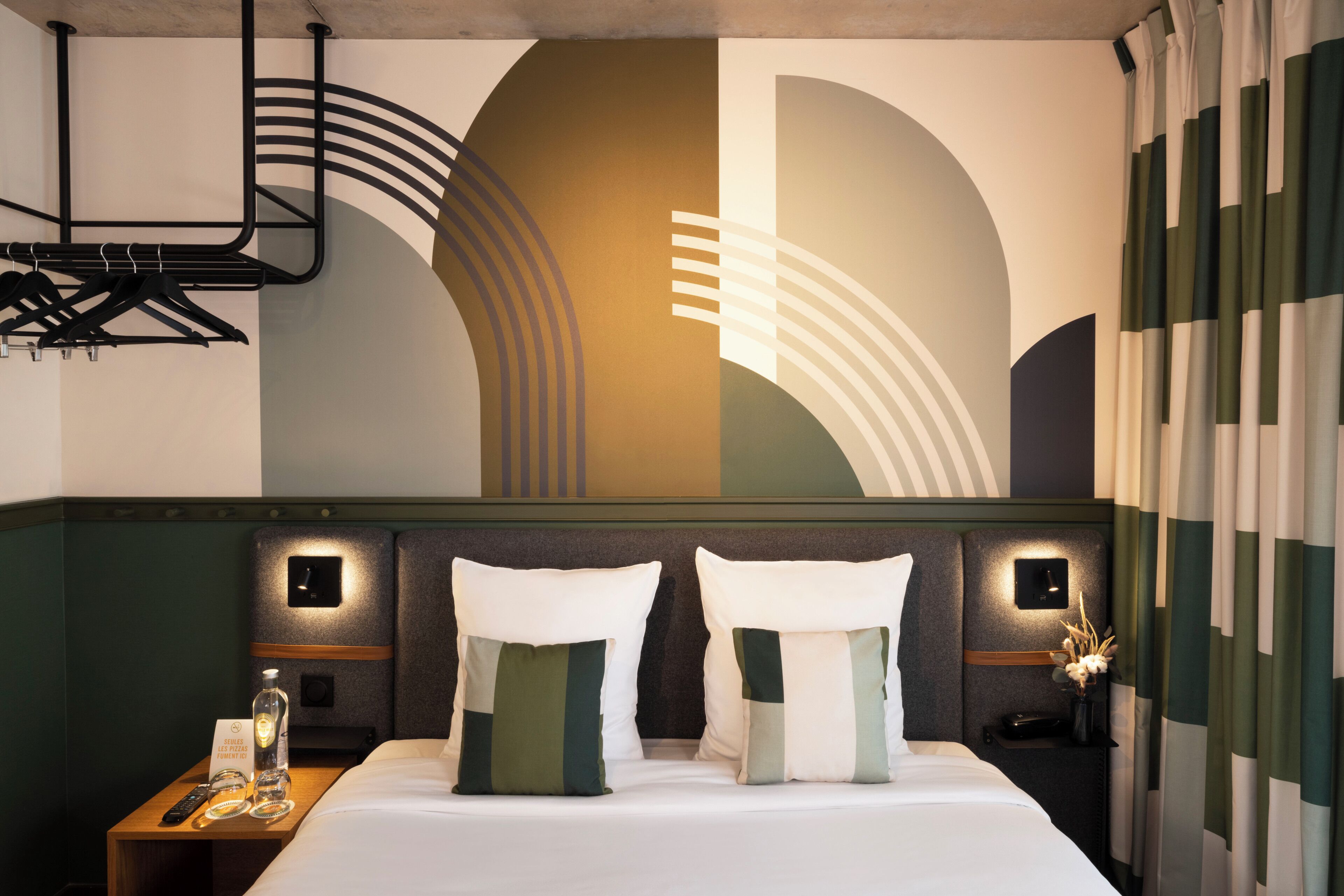Habitación de hotel con cama, dos almohadas, mesita de noche con control remoto, botella de vidrio y un pequeño florero con flores.