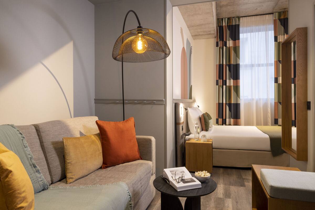 A modern hotel room with a sofa, a small table with a book and glasses, a bedside table with a lamp, a bed, and a window with curtains.