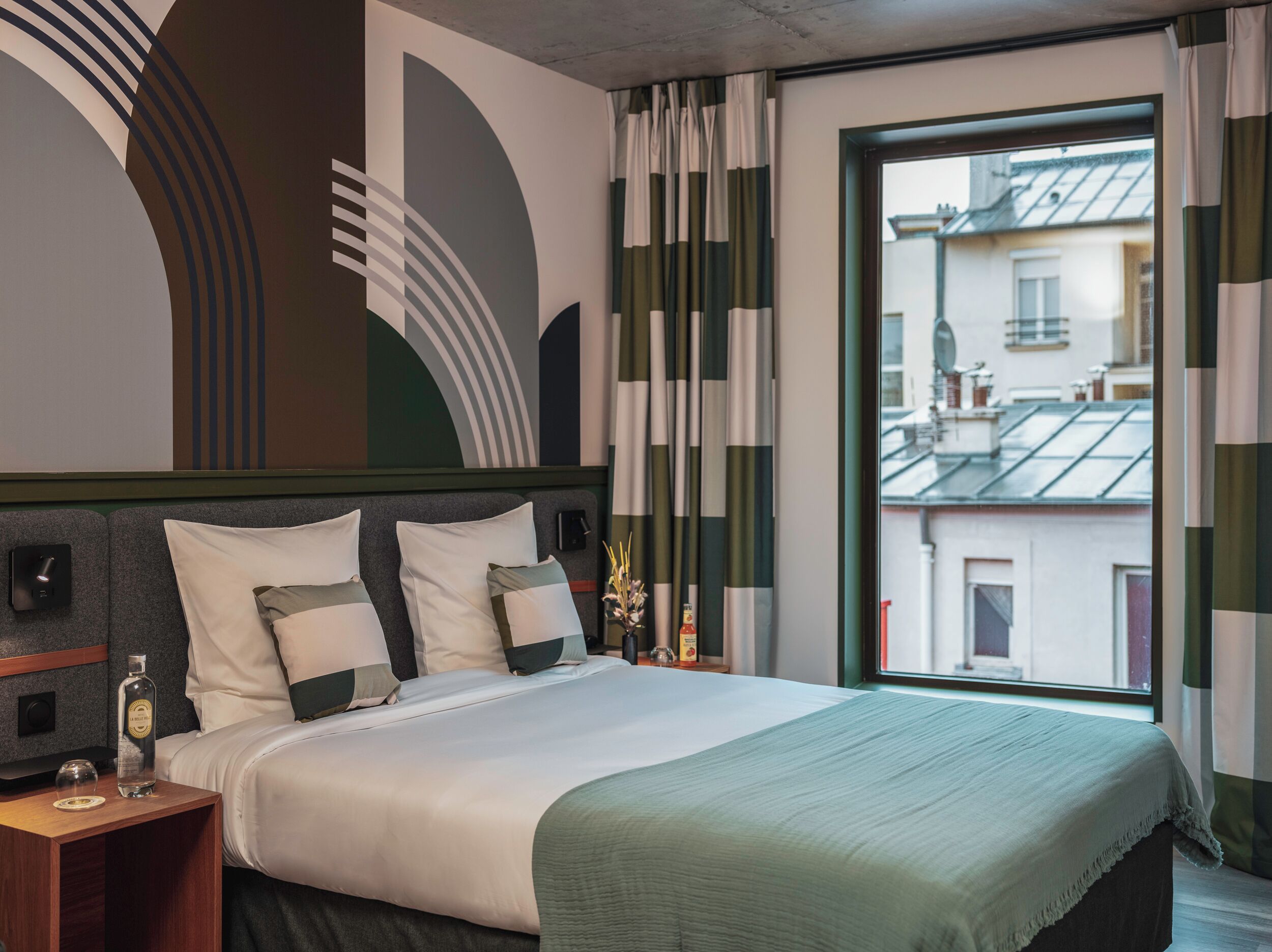 Quarto de hotel com uma cama grande, uma mesinha de cabeceira, uma garrafa de licor e vista para a cidade pela janela.
