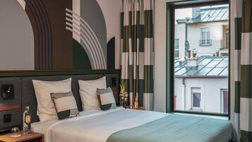 Quarto de hotel com uma cama grande, uma mesinha de cabeceira, uma garrafa de licor e vista para a cidade pela janela.