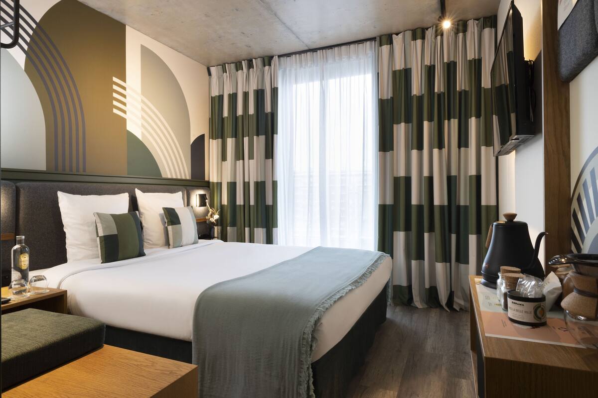 A hotel room with a large bed, a bedside table with a lamp, a coffee maker, and a TV on the wall.