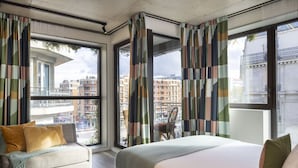 A modern hotel room with a large bed, a sofa, and a view of the cityscape through large windows. - La Belle Ville (Paris)