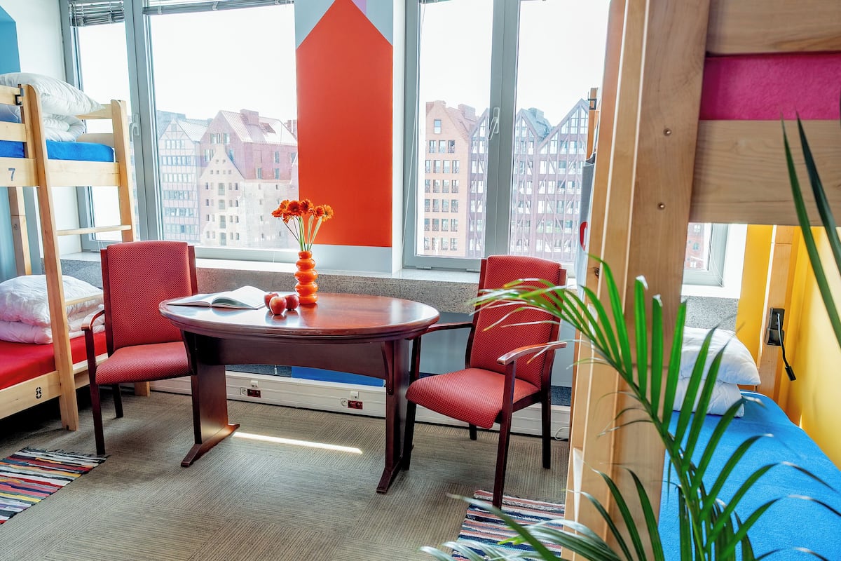 A room with a bunk bed, a dining table with chairs, a vase with flowers, and a large window with a view of buildings.