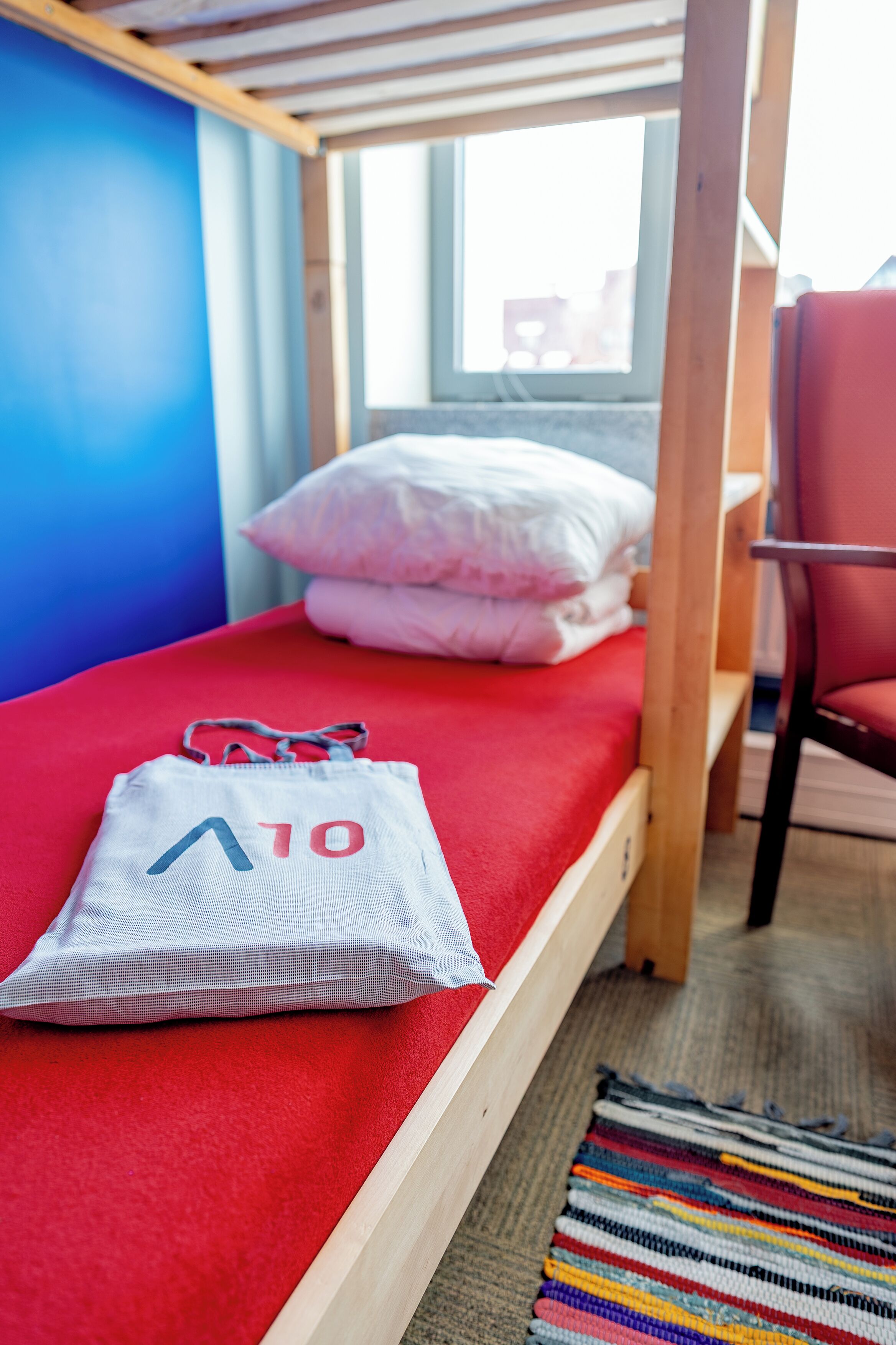 A bunk bed with a red mattress, a white pillow, and a white bag with red and blue text.