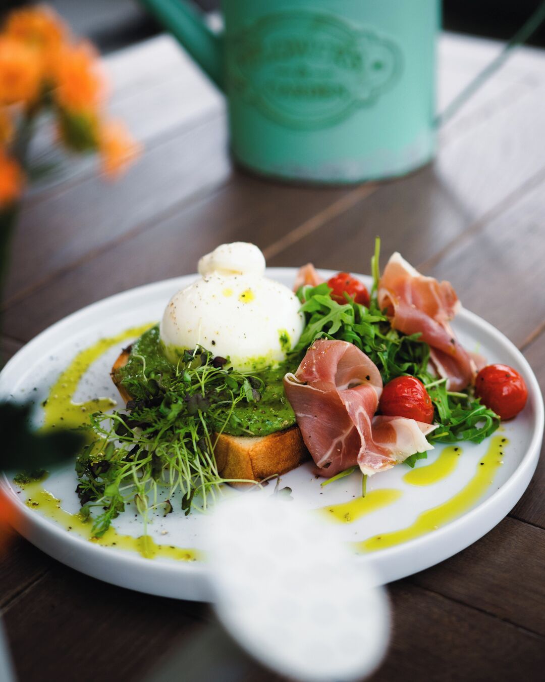 A plate of food with a poached egg, prosciutto, arugula, cherry tomatoes, and avocado on toast, served on a white plate with olive oil drizzles.