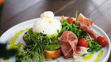 A plate of food with a poached egg, prosciutto, arugula, cherry tomatoes, and avocado on toast, served on a white plate with olive oil drizzles.