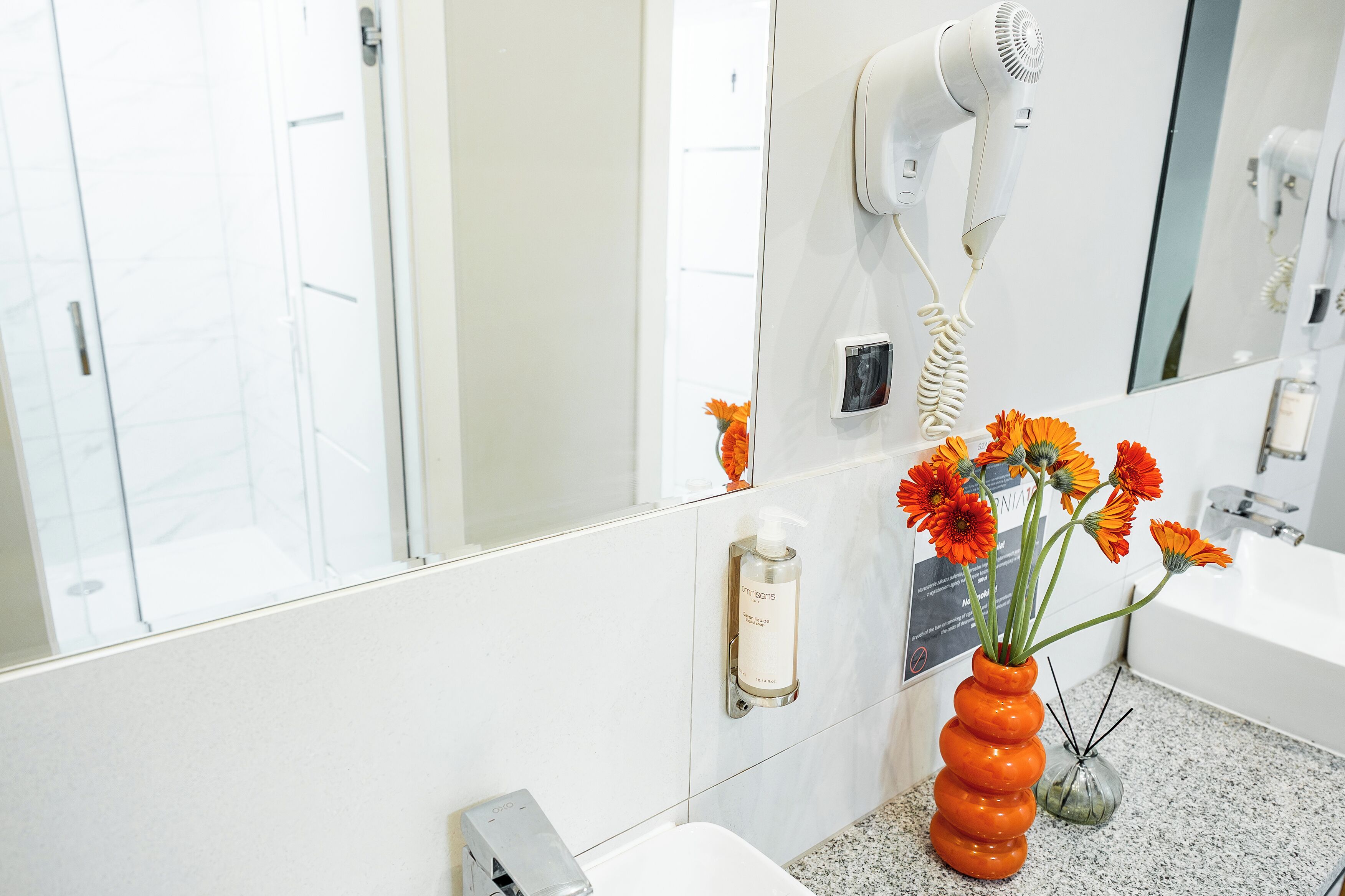 A bathroom with a white sink, a mirror, a hair dryer mounted on the wall, and a vase with orange flowers.