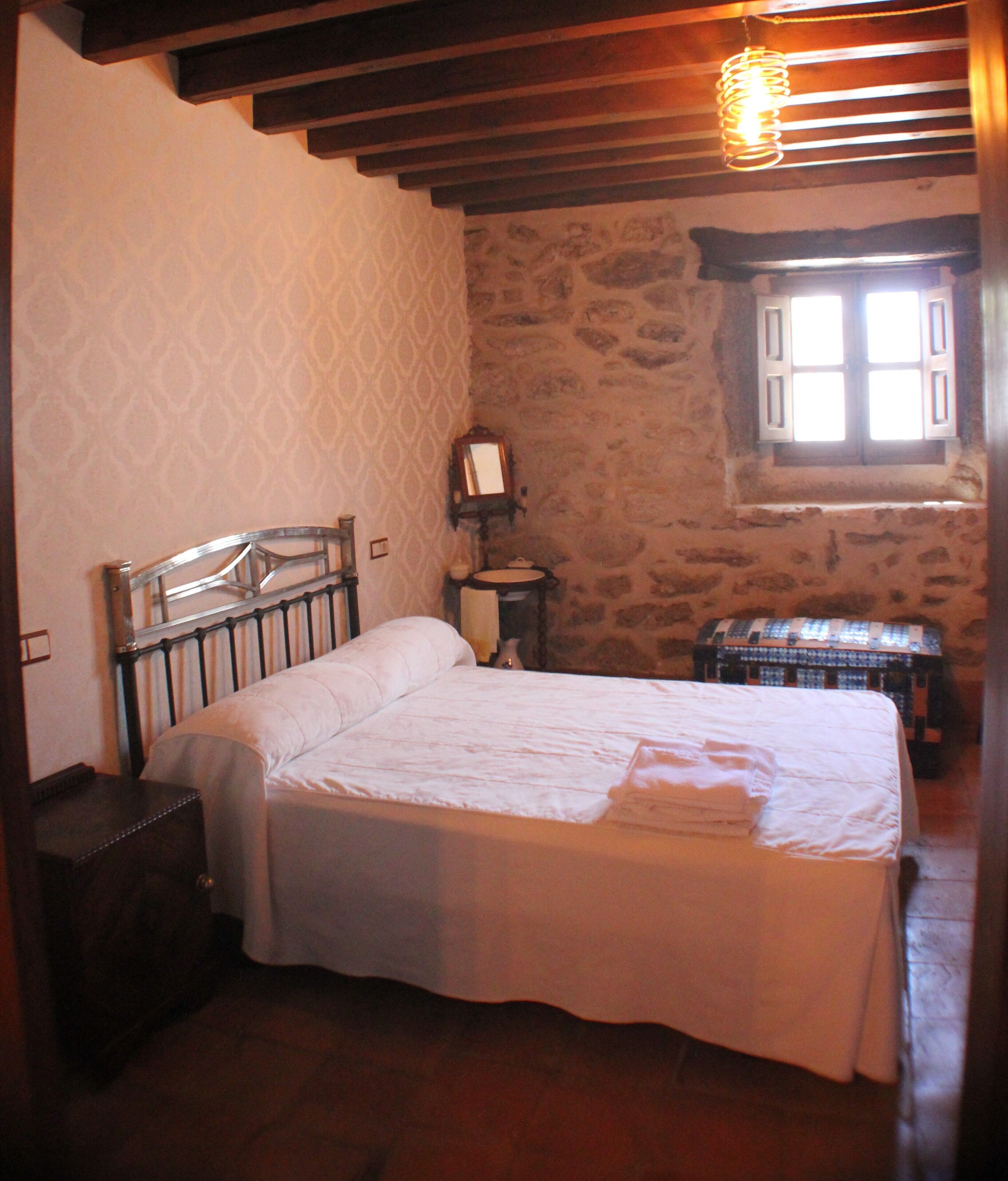A bedroom with a bed, a small bedside table, a mirror, a window, and a stone wall.