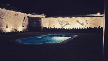 Vista noturna de um pátio com piscina, paredes de pedra e lanternas tradicionais.