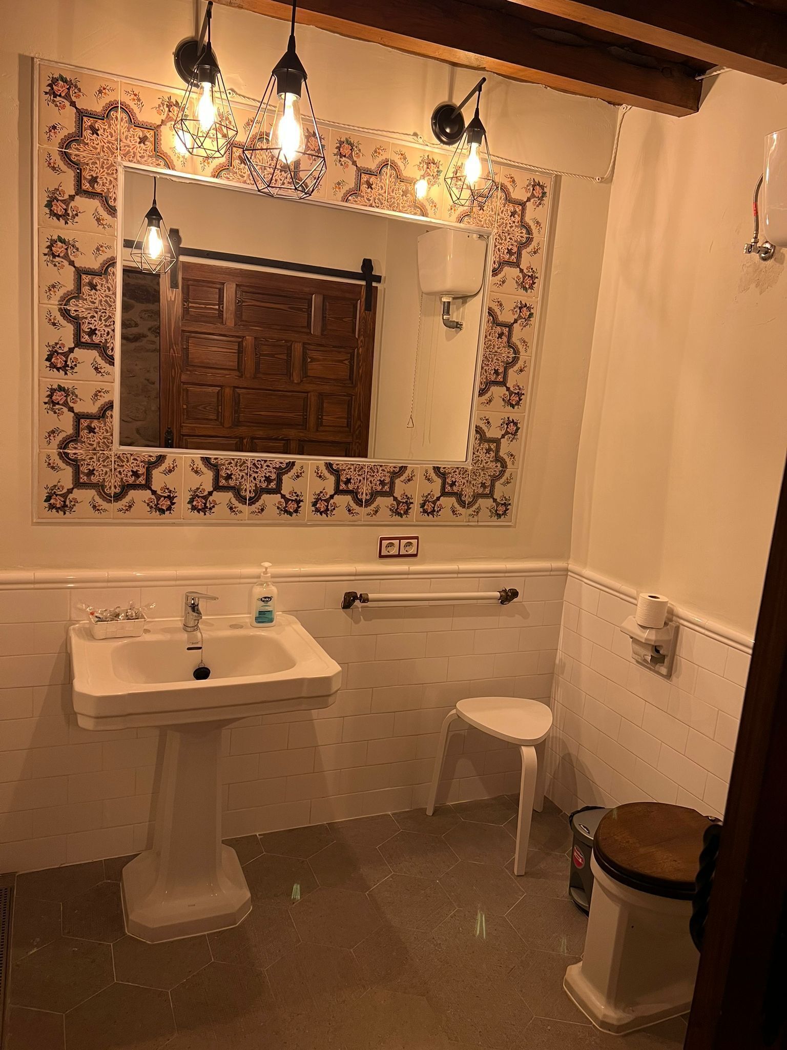 Une salle de bain avec un lavabo blanc, une porte en bois, un miroir orné d’un décor de carreaux et des toilettes.