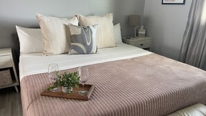 Ein ordentlich bezogenes Bett mit einer rosafarbenen Steude, ein Nachttisch mit einer Lampe und ein Tablett mit Gläsern und einer Pflanze.