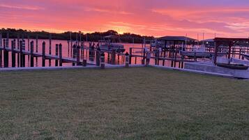 A grassy area with a dock extending into a body of water, surrounded by trees and a sunset sky.