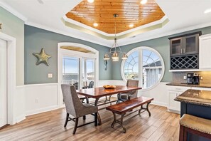 A dining area with a wooden table and chairs, a bench, a chandelier, and a view through large windows. - Beach Easy by Brunswick Vacation Rentals: Heated Pool & Spa, Tiki Bar, Elevator (Ocean Isle Beach)