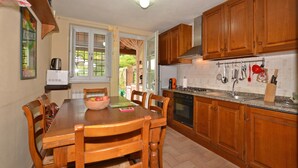 A kitchen with wooden cabinets, a dining table with chairs, a window with curtains, and a hanging pot rack. - Casa D'amelia (Camaiore)
