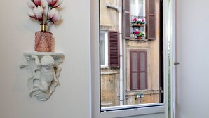 A window with a view of a building with brown shutters and a white wall with a decorative white shelf holding a vase with flowers. - Luxury apartment in Rione Monti (Roma)