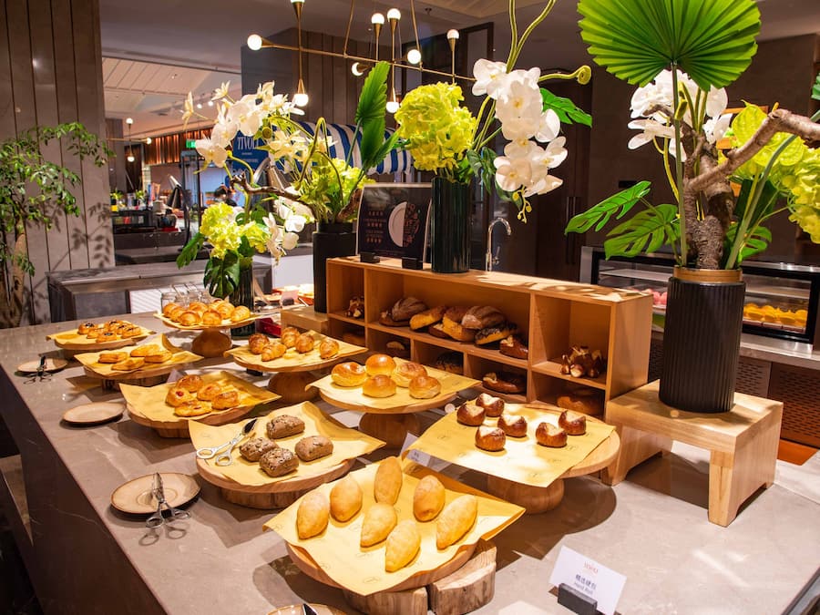 Vitrine de padaria com diversos produtos de panificação e um arranjo de plantas decorativo.