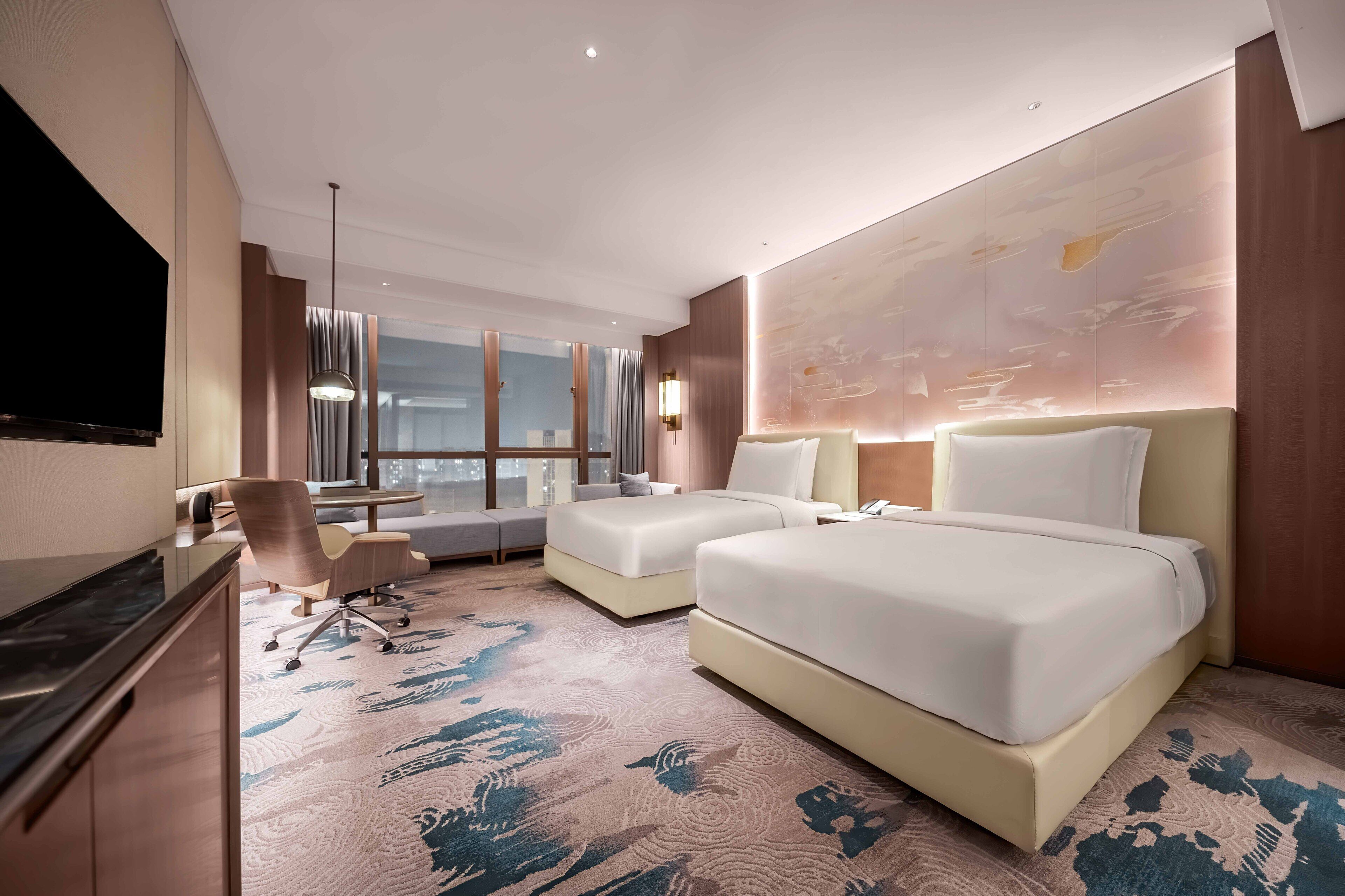 A modern hotel room with two beds, a desk, a sofa, and a large window.