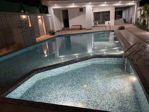 A swimming pool with mosaic tile flooring and metal railings, surrounded by a stone wall and a building with a covered patio area.