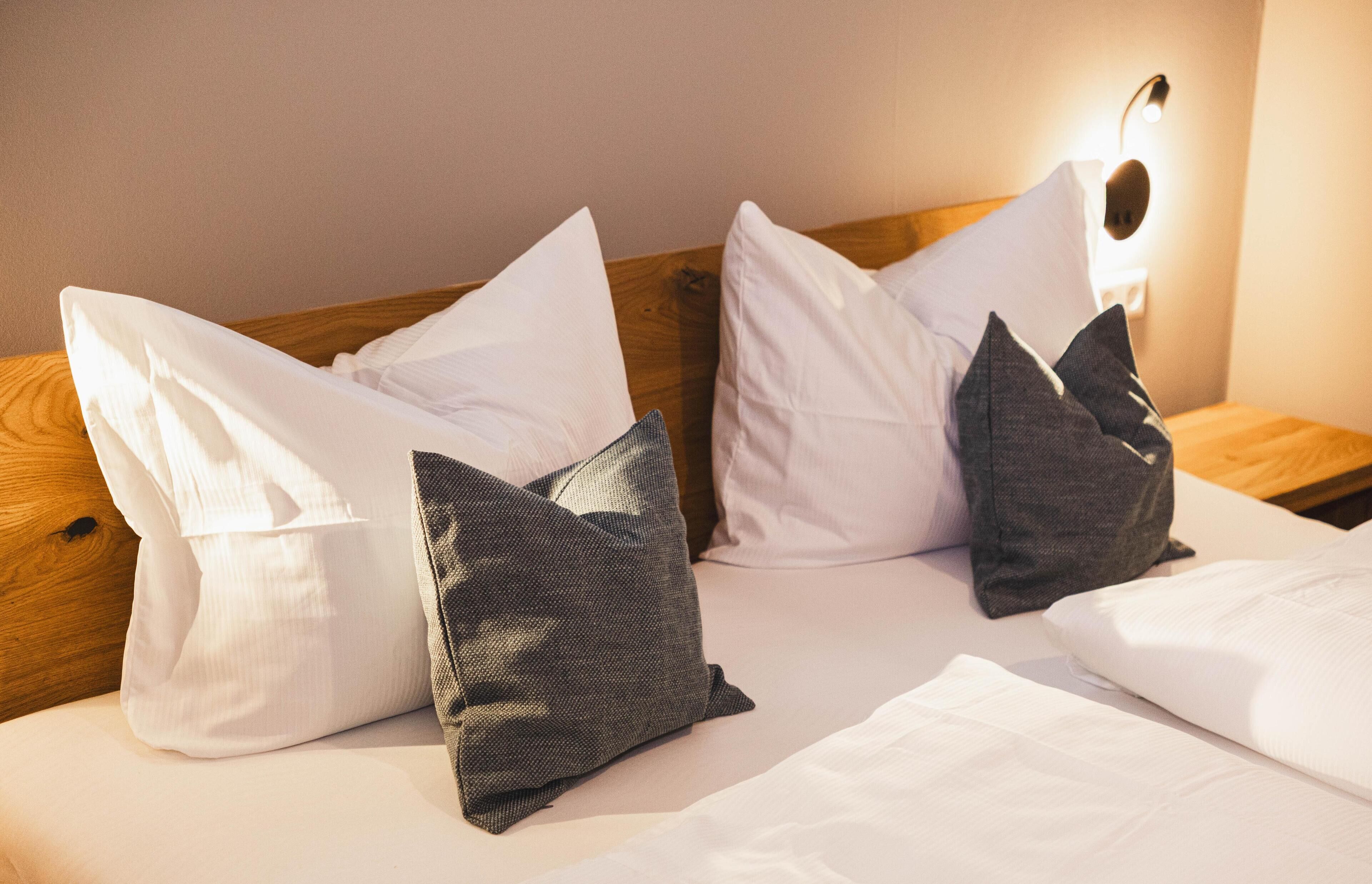 Un lit bien fait, avec des oreillers blancs et gris foncé, une tête de lit en bois et un luminaire fixé au mur.