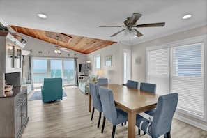 A spacious room with a wooden ceiling, a dining table with chairs, a sofa, and a balcony with a view.