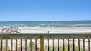 Vista de la playa con una terraza de madera y una persona sentada en la arena.