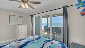 Un dormitorio con cama, ventilador de techo, cómoda, ventana con vista al océano y una silla.