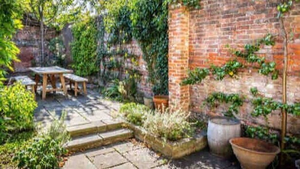 Un mur de briques recouvert de plantes grimpantes, une terrasse en pierre avec des marches et une table et des chaises en bois.