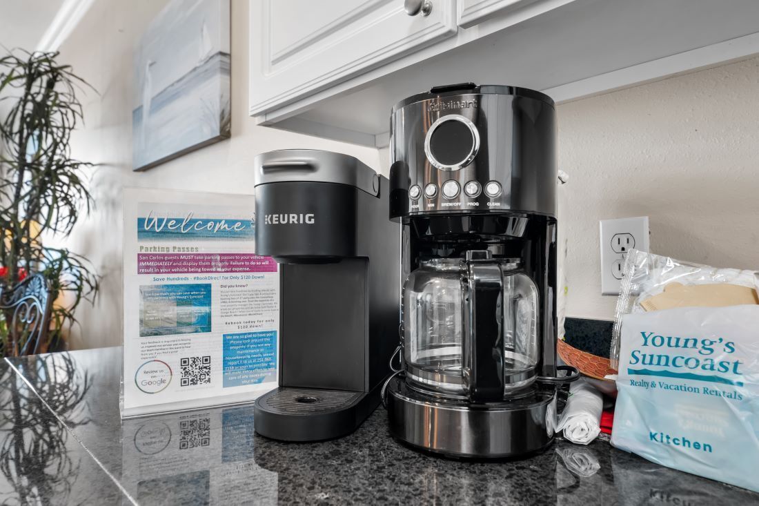 A kitchen counter with a Keurig coffee maker, a coffee pod, a Keurig branded mug, a Welcome sign, and a Young's Suncoast kitchen bag.