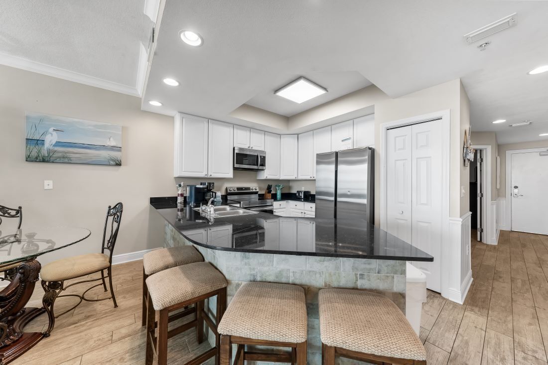 A modern kitchen with white cabinets, a black countertop, and stainless steel appliances. There is a dining area with a glass table and wicker chairs.