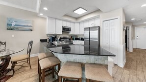 A modern kitchen with white cabinets, a black countertop, and stainless steel appliances. There is a dining area with a glass table and wicker chairs.