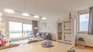 A modern living room with a sofa, coffee table, and a small ship model. There is a large window with curtains and a view of the outdoors. - Vacation apartment by the sea - LekkerNaarZee A203 (Callantsoog)