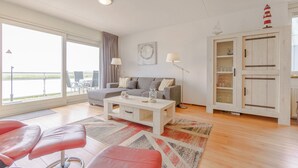A modern living room with a grey sofa, a white coffee table, and a red leather bench. There is a large window with a view of the sea, a lamp, and a cabinet with glass doors. - Vacation apartment by the sea - LekkerNaarZee A203 (Callantsoog)