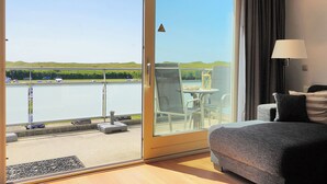 A room with a sliding glass door leading to a balcony overlooking a body of water and green hills. - Vacation apartment by the sea - LekkerNaarZee A203 (Callantsoog)
