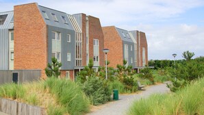 A modern building with a brick facade, multiple windows, and a pathway leading to a bench. - Vacation apartment by the sea - LekkerNaarZee A203 (Callantsoog)