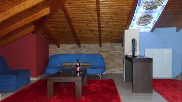 A cozy attic room with a sloped ceiling, a blue sofa, a small table with a bottle and glasses, a red rug, and a television on a stand.