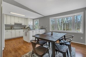A kitchen with white cabinets, a wooden dining table with four chairs, and large windows with a view of trees.