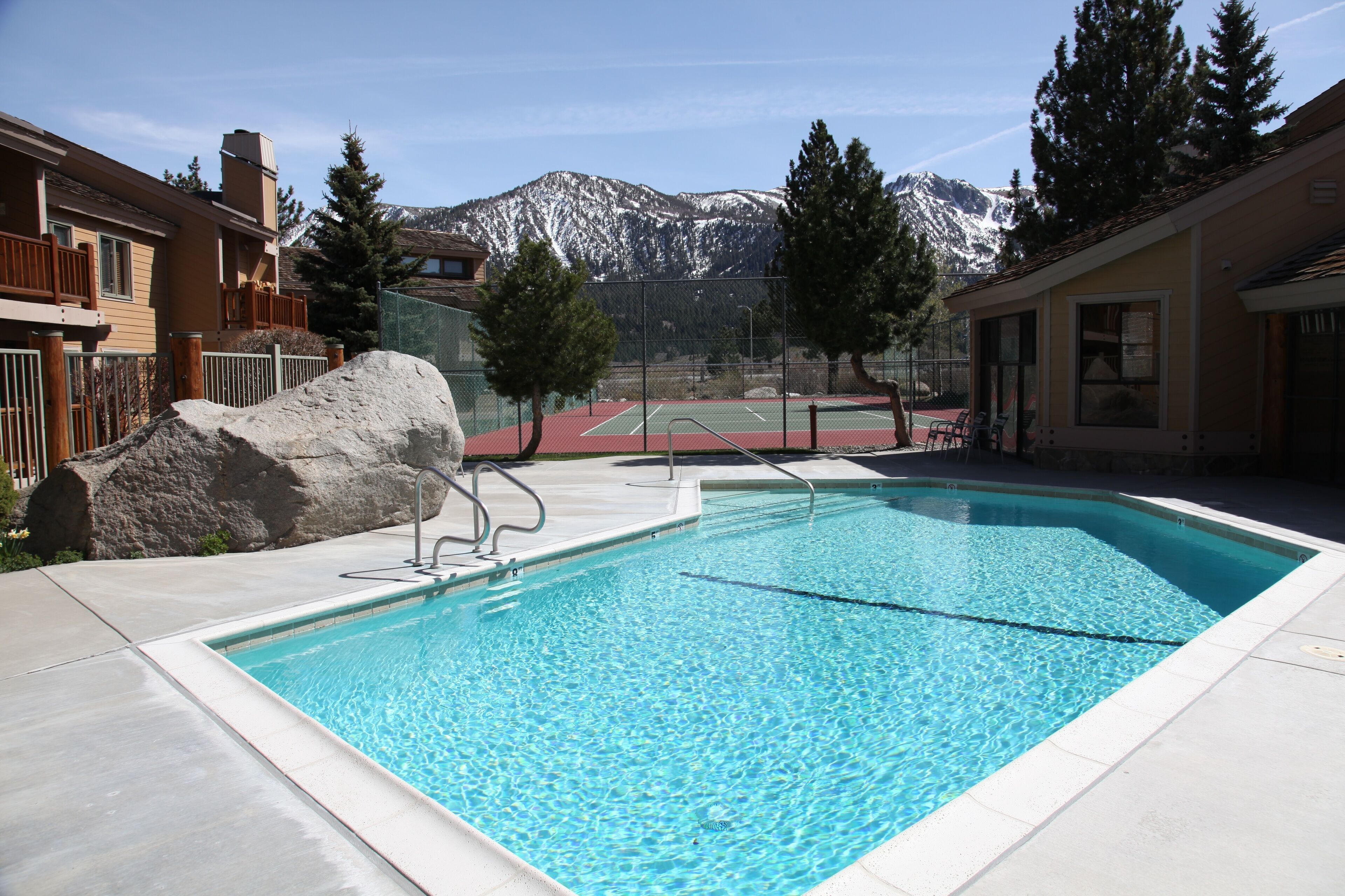 Una piscina con vistas a montañas nevadas, una cancha de tenis y una gran roca.