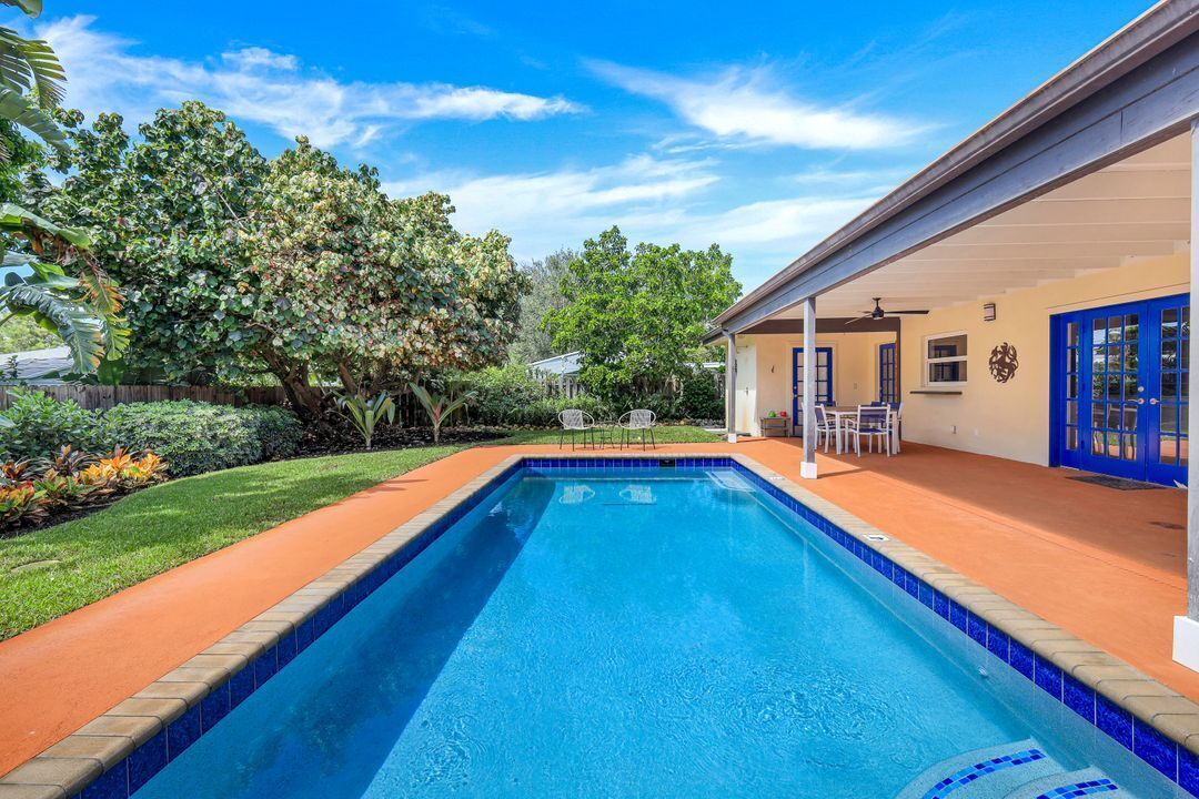 A rectangular swimming pool with a tiled edge, surrounded by a wooden deck and a covered patio area with chairs.