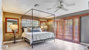 Un lit à baldaquin avec un ventilateur de plafond, une fenêtre avec des stores et une table de chevet avec une lampe.