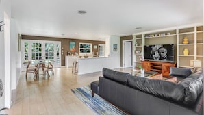 Un vaste espace de vie comprenant un canapé en cuir noir, une table basse en verre et un téléviseur. On y trouve également un coin repas avec des chaises en bois et une table en verre. La pièce présente un design moderne, avec des murs aux couleurs claires et un plancher en bois.