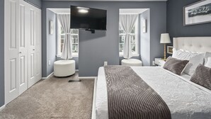 Una camera da letto con un letto, una TV a parete, due finestre con tende, un comodino con una lampada e un quadro appeso al muro.