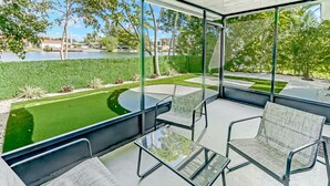 A modern outdoor patio with glass walls, a seating area, and a view of a well-maintained garden and water body. - Hallandale 1037 | Lakeside View: 15KM to Stadium & Near Beach (Hallandale Beach)