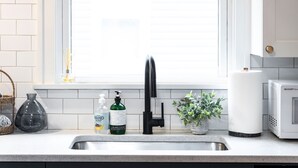 A kitchen with a white countertop, a stainless steel sink, a black faucet, a window with blinds, a potted plant, a microwave, and a basket. - City Boho ✦ New Kitchen ✦ King Bed (Bexley)