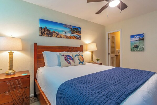 A bedroom with a wooden headboard, two bedside lamps, a blue quilted bedspread, and two wall-mounted paintings depicting coastal scenes.
