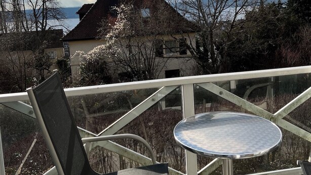 Ein Balkon mit einem Metallstuhl und einem kleinen runden Tisch, mit Blick auf ein Haus und Bäume.