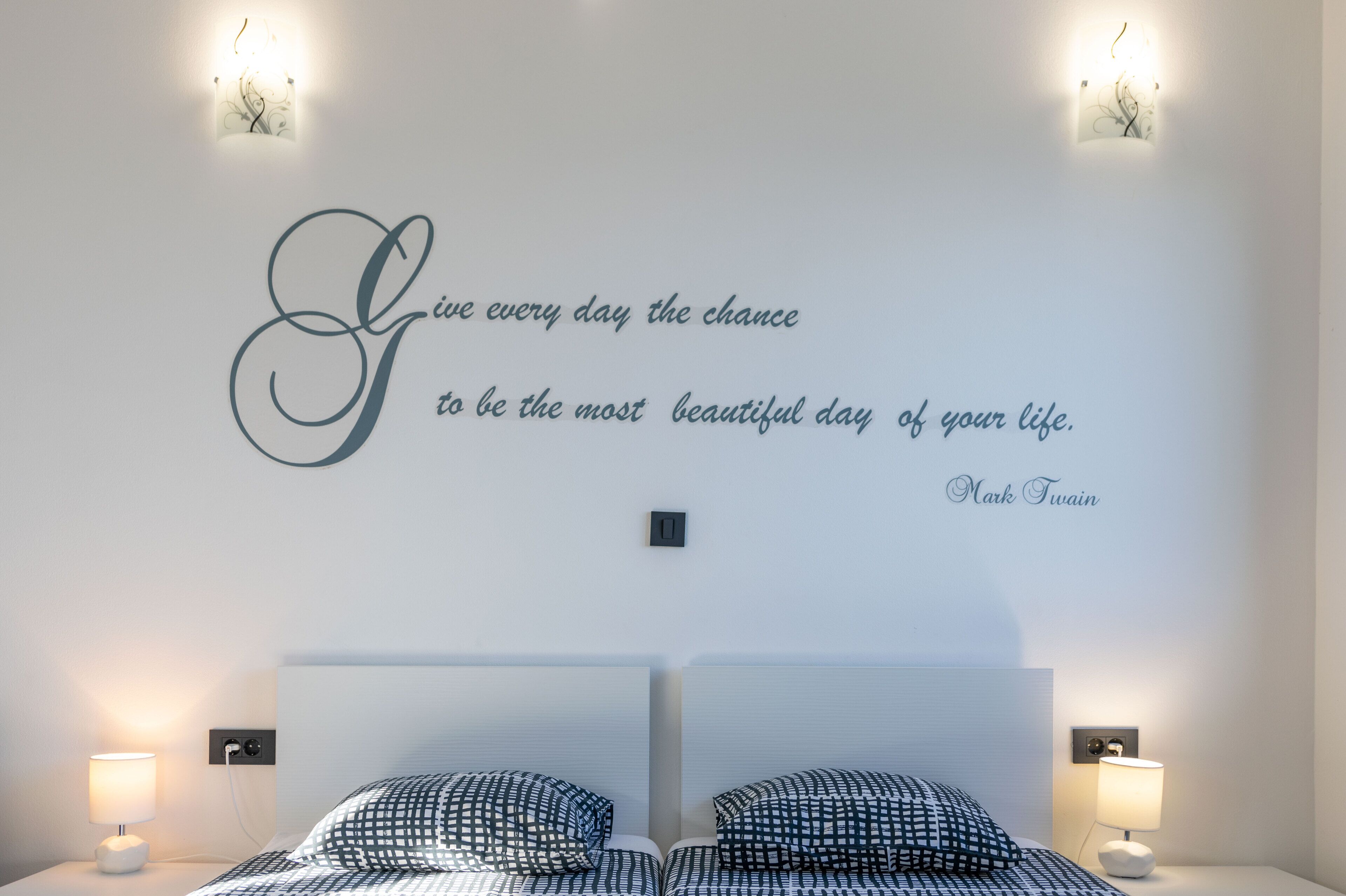 A bedroom with a bed, two bedside lamps, a wall-mounted light fixture, and a motivational quote.