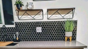 Una cocina con armarios blancos, un salpicadero hexagonal blanco y negro, y dos estantes montados en la pared con plantas y objetos decorativos.