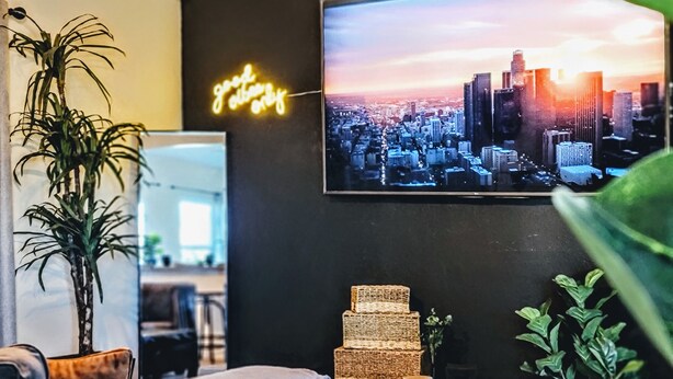 Una sala moderna con televisión mostrando un paisaje urbano, un letrero de neón y una pila de cestas tejidas.