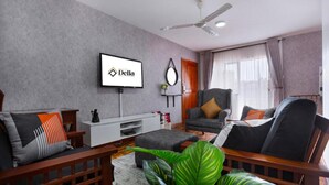 A modern living room with a grey sofa set, a wooden coffee table, and a grey armchair. A flat-screen TV mounted on the wall displays the logo "Della". - Delight Homes by Della (Nairobi)