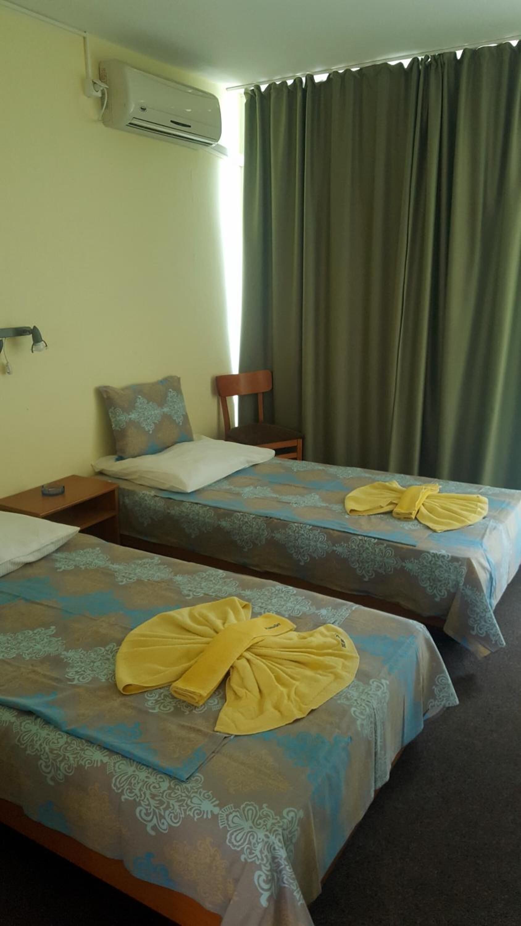 Quarto de hotel com duas camas, uma cadeira, uma mesa de cabeceira, ar condicionado e uma janela com cortinas.